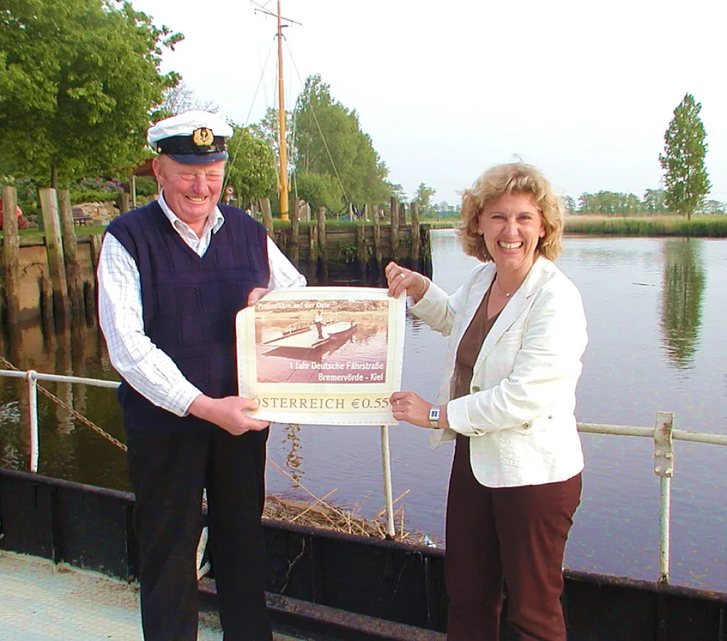 Helmut Plate ist gerührt über die neue österreichische Sondermarke, die seine Flussfähre zeigt. Karin Harms (AG Osteland) präsentiert dem Fährmann das Postwertzeichen mitten auf der Oste.
