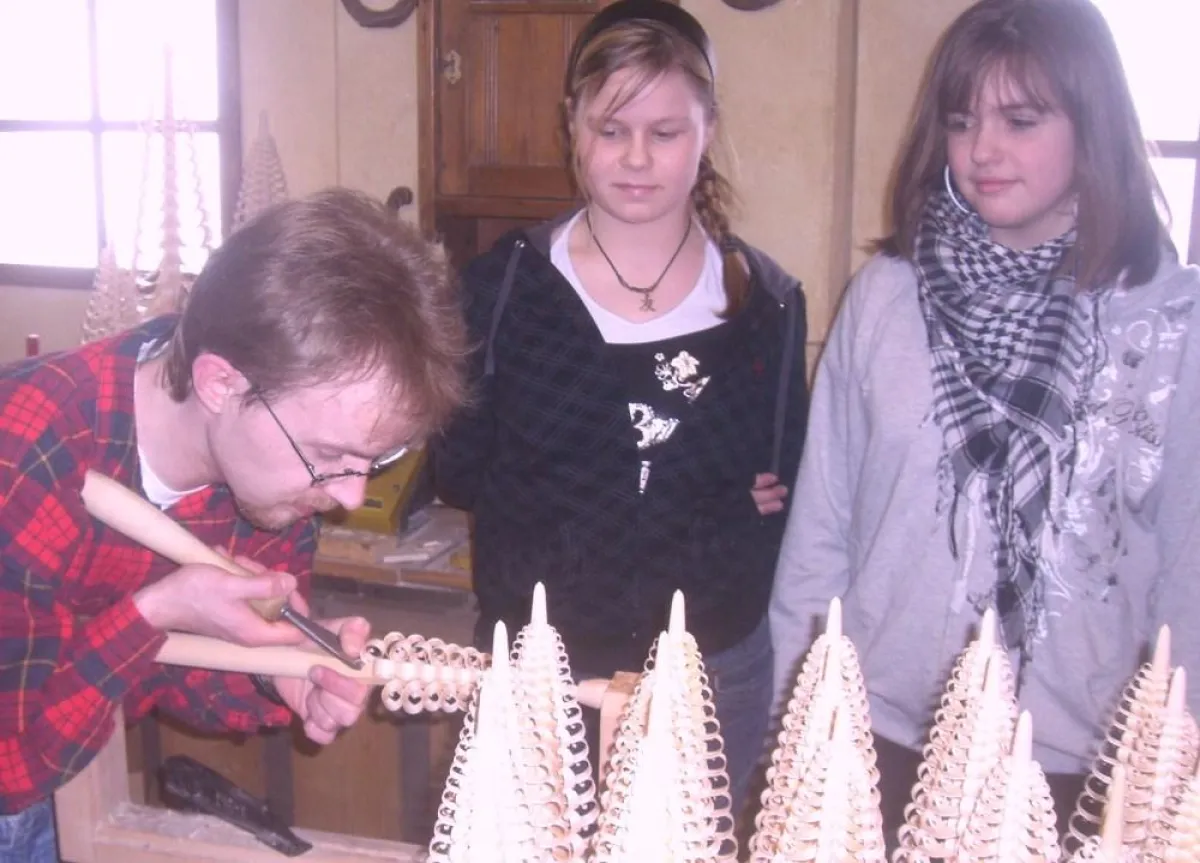 Anne ( 13 ) und Katharina ( 12 ) schauen in der Schauwerkstatt im Stracoland in Colmnitz / Sachsen einem Spanbaumstecher bei der Erstellung eines traditionell erzgebirgischen Spanbaums zu.