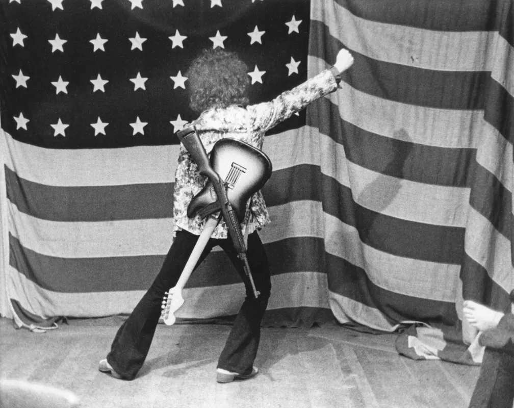 Gitarrist Wayne Kramer von der Gruppe MC5 posiert vor einer amerikanischen Flagge im Jahr 1969 in E (© Leni Sinclair)