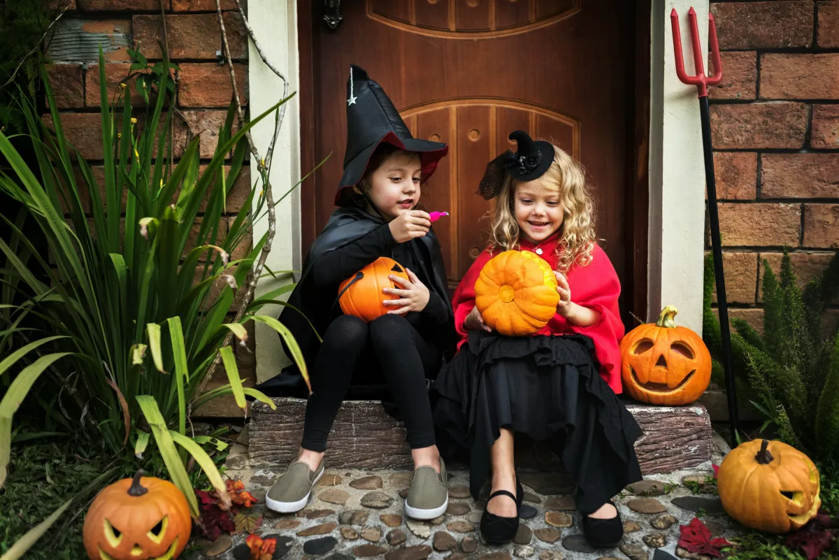 Zwei Kinder im Halloweenkostüm sitzen lachend vor einer Haustür mit Kürbissen (© Curated Lifestyle by Unsplash)