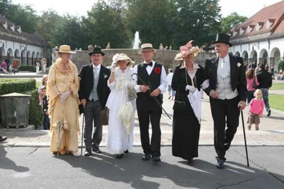 Damen mit aufsehenerregenden Hüten und galante Herren mit Frack und Zylinder flanieren im Sprudelhof