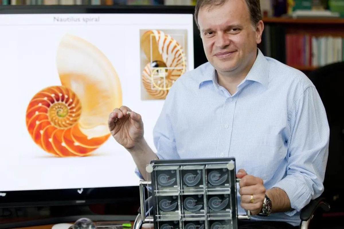 Professor Dr. Dr. Andrej Kral, der das Modell eines menschlichen Innenohres zeigt. Im Hintergrund eine Nautilus-Schale, im Vordergrund Vergrößerungen verschiedener Innenohre. (