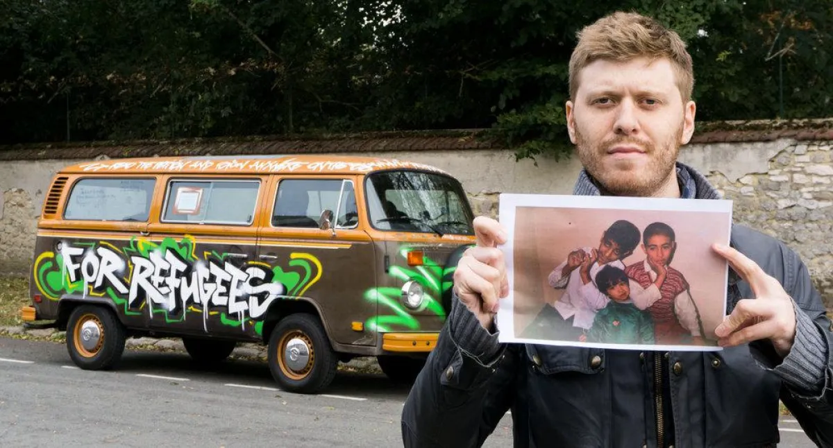 Grigorij Richters hält ein Bild auf dem drei Flüchtlingskinder zu sehen sind. Mit VW Bus im Hintergr